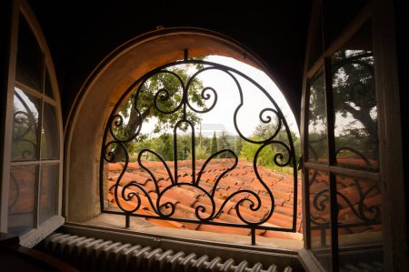 Photo for An open arch window, equipped with forged metalwork bars in form of round arabesques, overlooking a tiled roof and a park with verdant trees, seen from the interior of a traditional French manor house - Royalty Free Image