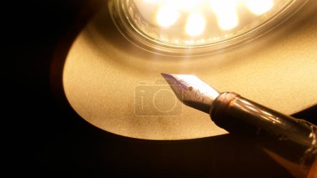 A LED lamp in the domed form of a cupola radiating yellow light illuminates an old metal fountain pen that features a worked golden nib with stains of blue ink, in warm colors with a dark background 