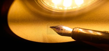 Foto de Una lámpara led en forma de cúpula de una cúpula que irradia luz amarilla ilumina una vieja pluma estilográfica de metal que cuenta con un plumín dorado trabajado con manchas de tinta azul, en colores cálidos con un fondo oscuro - Imagen libre de derechos