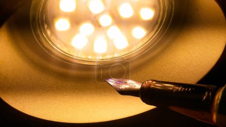 Foto de Una lámpara led en forma de cúpula de una cúpula que irradia luz amarilla ilumina una vieja pluma estilográfica de metal que cuenta con un plumín dorado trabajado con manchas de tinta azul, en colores cálidos con un fondo oscuro - Imagen libre de derechos