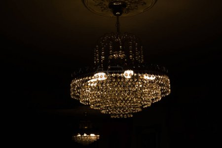 Téléchargez les photos : Lustre en cristal luxueux, suspendu au plafond d'un salon, et émettant de la lumière jaune à travers des lampes à ampoule électrique, au crépuscule, dans un environnement sombre - en image libre de droit