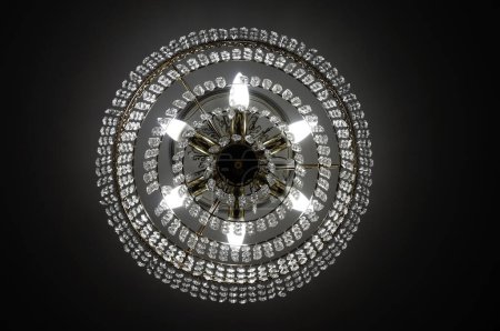 Photo for Luxurious crystal chandelier, hanging from the ceiling of a living room, and emitting yellow light through electric bulb lamps, in a the twilight, in a dark environment - Royalty Free Image