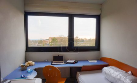 Foto de Reims, Francia - Feb. 2021 - Una pequeña y moderna habitación de estudiantes en Evariste Galois CROUS Residence, en el campus URCA Moulin de la Housse: hay un ordenador en un escritorio, frente a un gran ventanal - Imagen libre de derechos