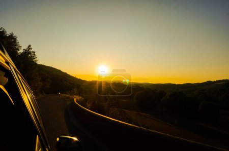Téléchargez les photos : Grand paysage rural flou par un coucher de soleil aveuglant à l'horizon sur la ligne d'horizon du massif des montagnes centrales de l'Aveyron, dans le sud de la France, avec une route courbe traversant les champs et les forêts - en image libre de droit