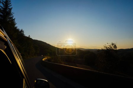 Foto de Gran paisaje rural borroso por una puesta de sol cegadora en el horizonte sobre el horizonte de las montañas Macizo Central en Aveyron, sur de Francia, con una carretera curva que cruza campos y bosques - Imagen libre de derechos
