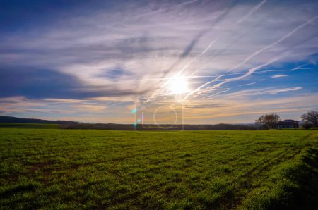 Téléchargez les photos : Beau paysage rural en France avec un champ vert paisible, une fuite de lumière et la lumière du soleil à travers les nuages et les traînées d'avion au début du coucher du soleil - en image libre de droit