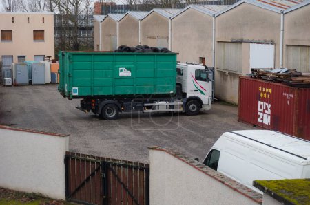 Foto de Occitanie, Francia - Feb. 2021 - Un camión Volvo de la empresa francesa de reciclaje Aliapur, cargando un cubo desmontable verde cargado con neumáticos de goma usados, en el patio de un centro de almacenamiento - Imagen libre de derechos
