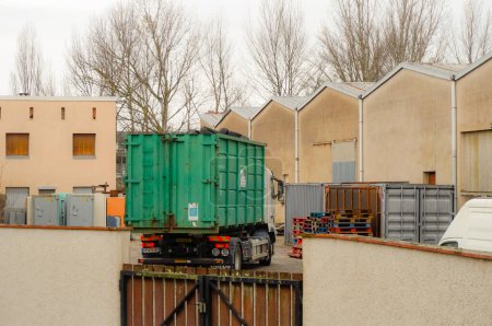 Foto de Occitanie, Francia - Feb. 2021 - Un camión Volvo de la empresa francesa de reciclaje Aliapur, cargando un cubo desmontable verde cargado con neumáticos de goma usados, en el patio de un centro de almacenamiento - Imagen libre de derechos