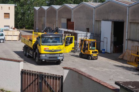 Téléchargez les photos : Albi, France - Juin 2021 - Un camion Renault jaune (Groupe Volvo Trucks), équipé d'un godet amovible et d'une flèche hydraulique, et d'un chariot élévateur à fourche, dans la cour d'une usine, devant les hangars - en image libre de droit