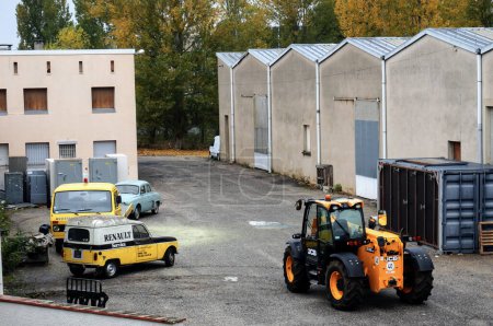 Photo for Albi, France - Dec. 2018 - A JCB telescopic handler, old vehicles (a Renault minivan, a truck and a sedan car) and a shipping container in front of the warehouse of a storage facility - Royalty Free Image
