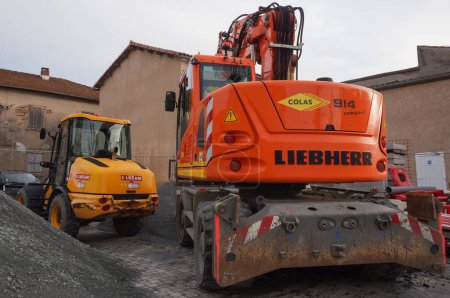 Foto de Tarn, Francia - Feb. 2021 - Dos máquinas de construcción de ruedas en un lugar de trabajo: una excavadora Liebherr naranja, operada por Colas, en primera línea y una cargadora articulada amarilla, alquilada por Loxam - Imagen libre de derechos