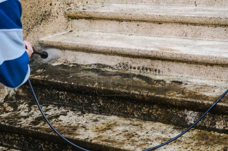 Téléchargez les photos : Entretien extérieur : un ouvrier nettoie et démousse un escalier en pierre extérieur sale avec la lance d'un laveur haute pression, tandis que la mauvaise eau, chargée de mousse, trébuche sur les marches - en image libre de droit