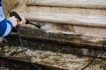 Mantenimiento de la casa: un trabajador manual limpia y deshace una escalera de piedra exterior sucia con la lanza de una arandela de alta presión, mientras que el agua mala, cargada de musgo, baja por las escaleras
