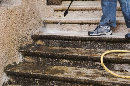 Photo for House maintenance : a manual worker wearing torn blue trousers cleans and defoams a dirty exterior stone staircase with the lance of a high-pressure washer, while the bad water trikles down the steps - Royalty Free Image