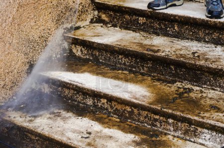Photo for Outdoor maintenance : a manual worker cleans and defoams a dirty exterior stone staircase with the lance of a high-pressure washer, while the bad water, charged with moss, trikles down the steps - Royalty Free Image