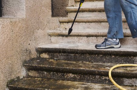 Photo for House maintenance : a manual worker wearing torn blue trousers cleans and defoams a dirty exterior stone staircase with the lance of a high-pressure washer, while the bad water trikles down the steps - Royalty Free Image