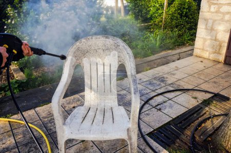 Téléchargez les photos : Activités de plein air au printemps : une chaise de jardin en plastique blanc sale est nettoyée avec la buse d'eau d'un laveur haute pression, qui met en évidence l'effet avant et après entre les zones teintées et nettoyées - en image libre de droit