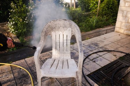 Téléchargez les photos : Activités de plein air printanières : une chaise de jardin en plastique blanc sale est nettoyée avec la buse d'eau d'un laveur haute pression, qui met en évidence l'effet avant et après entre les zones teintées et nettoyées - en image libre de droit