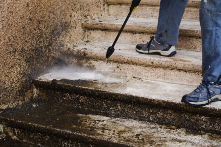 Hauswartung: Ein Arbeiter in zerrissener blauer Hose säubert und schäumt eine schmutzige Steintreppe mit der Lanze eines Hochdruckreinigers, während das schlechte Wasser die Treppe hinunterrieselt