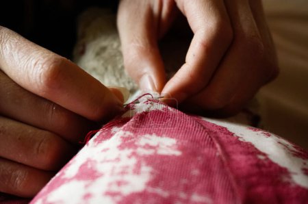 Photo for Closeup on the hands of a female couturier skillfully handling a stitching neddle and repairing the red discolored fabric of an old damaged stuffed toy animal - Royalty Free Image