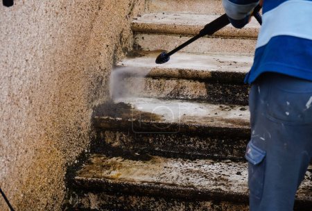 Trabajo al aire libre: un oficial de mantenimiento limpia y deshace una escalera de piedra exterior sucia con la lanza de una arandela de alta presión, mientras que el agua mala, cargada de musgo, baja por las escaleras