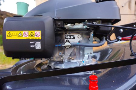 Foto de Tarn, Francia - Mayo 2020 - Detalle de la unidad de motor de una cortadora de césped de combustión alimentada por el fabricante japonés Honda, con la válvula de combustible en la manguera de entrada, el acelerador y los cables de embrague en el motor - Imagen libre de derechos