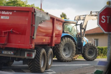 Téléchargez les photos : Tarn, France - Août 2021 - Un tracteur agricole bleu avec une chargeuse frontale remorquant une remorque, prend un virage à un carrefour sur une route rurale, dans une zone rurale du Tarn, dans le sud-ouest de la France - en image libre de droit