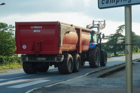 Téléchargez les photos : Tarn, France - Août 2021 - Un tracteur agricole bleu avec une chargeuse frontale remorquant une remorque rouge Maitre Atlas à deux roues sur une route rurale, dans une zone rurale du Tarn, dans le sud-ouest de la France - en image libre de droit