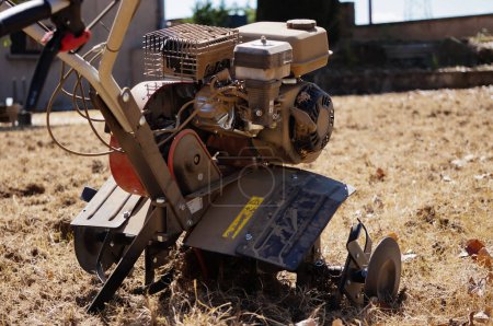Téléchargez les photos : Un motoculteur rotatif recouvert de poussière en action, utilisé dans un jardin pour labourer un sol sec et sablonneux ; ce puissant modèle assemblé en France dispose d'un moteur à deux temps fabriqué en Chine, et de six dents en acier - en image libre de droit