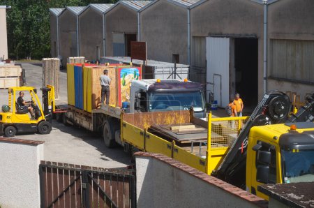 Téléchargez les photos : Albi, France - Juin 2021 - Activité dans un centre de stockage : quelques travailleurs déchargent le fret de deux camions à flèche jaune au moyen d'un chariot élévateur à fourche, devant les hangars de l'installation industrielle - en image libre de droit