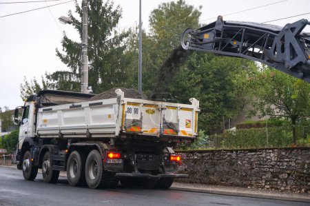 Photo for Albi, France - Aug. 2021 - A white dump truck on the street receives in its tipper, from a conveyor belt, the old pavement removed by a cold milling machine, during works of road maintenance - Royalty Free Image