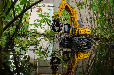 Foto de Occitanie, Francia - Mayo 2020 - Modelo a escala (juguete infantil) de una excavadora de orugas equipada con una garra hidráulica unida al brazo mecánico, que opera en el agua en un paisaje pantanoso salvaje - Imagen libre de derechos