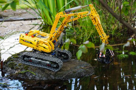 Foto de Occitanie, Francia - Mayo 2020 - Modelo a escala (juguete infantil) de una excavadora de orugas equipada con una garra hidráulica unida al brazo mecánico, que opera en el agua en un paisaje pantanoso salvaje - Imagen libre de derechos