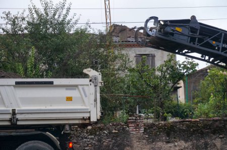 Téléchargez les photos : Albi, France - 1er août 2021 - À droite, de l'asphalte arraché de la route est versé dans le seau d'un camion benne, à gauche, par la bande transporteuse d'une fraiseuse, sur un chantier de construction - en image libre de droit