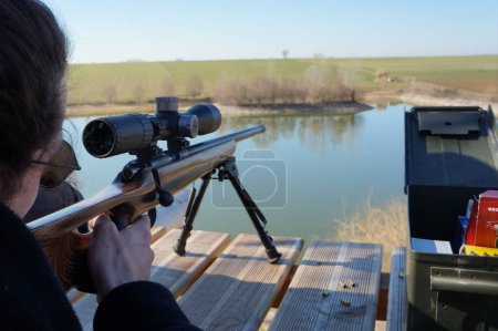 Photo for Occitanie, France - Feb. 2023 - A marksman aims at his target beyond a pond, in the fields, pointing thanks to a riflescope mounted on his Czech CZ 457 "Winchester" carbin, in a vast firing range - Royalty Free Image