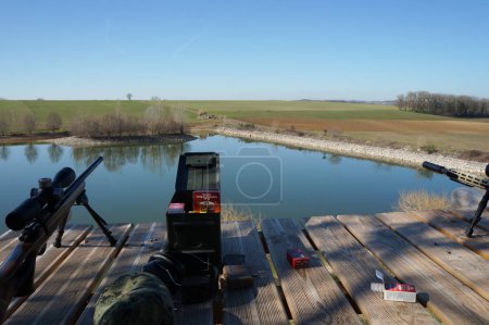Photo for Occitanie, France - Feb. 2023 - A wooden shooting platform, with two CZ 457 and Ruger Precision Rimfire "Winchester" carbins, and ammunition boxes, dominates a pond and fields in a vast firing range - Royalty Free Image