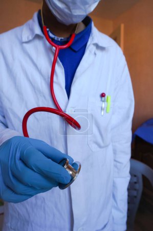 Téléchargez les photos : Albi, France - Avril 2020- Un médecin portant une blouse blanche, un masque et des gants pour limiter la contagion au sein de l'épidémie de coronavirus, détient un stéthoscope pour une auscultation (photo mise en scène) - en image libre de droit