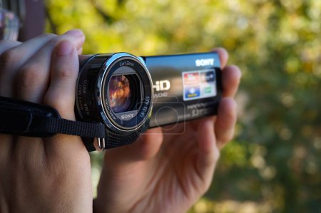 Téléchargez les photos : Occitanie, France - Novembre 2020 - Les deux mains du cameraman tenant un petit caméscope numérique, conçu par le fabricant japonais Sony, filmant une vidéo haute définition dans un jardin, en arrière-plan bokeh - en image libre de droit