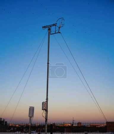Foto de Poste de acero estabilizado por cables de la estación meteorológica situada en la parte superior del aparcamiento Carmes, con sensores para mediciones meteorológicas y control de la contaminación atmosférica, en un fondo de cielo azul al amanecer - Imagen libre de derechos