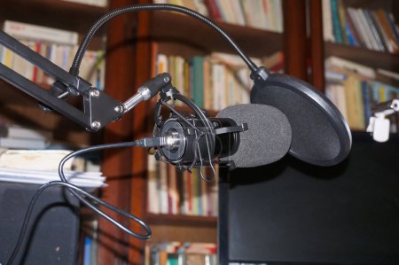 Foto de Micrófono de grabación profesional de gama alta equipado con un montaje, una protección de doble parabrisas de espuma negra, conectado con un cable USB e instalado en una biblioteca de la casa, delante de las estanterías - Imagen libre de derechos