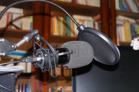 Foto de Micrófono de grabación profesional de gama alta equipado con un montaje, una protección de doble parabrisas de espuma negra, conectado con un cable USB e instalado en una biblioteca de la casa, delante de las estanterías - Imagen libre de derechos