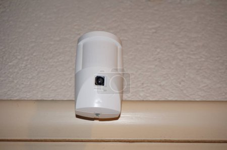 Téléchargez les photos : Un détecteur de mouvement équipé d'une caméra de surveillance, dans le cadre d'un système d'alarme et de télésurveillance dans une maison connectée, fixé sur un cadre de porte - en image libre de droit