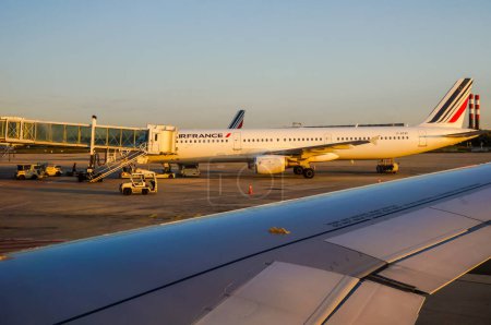 Foto de Aeropuerto de París, Francia - junio 2019- Avión Airbus A320 bimotor de media distancia de la aerolínea Air France, conectado a la puerta de la terminal a través de un puente de embarque, con tractores de aviones y de equipaje - Imagen libre de derechos
