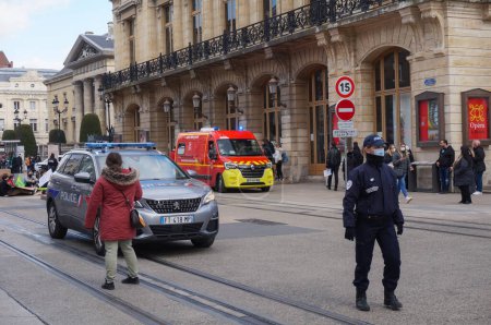 Foto de Reims, Francia - Marzo 2021 - Una ambulancia médica roja de la brigada de bomberos se dirige a la calle Vesle, en el centro, mientras que una oficial de policía femenina, frente a un Peugeot 5008, controla el tráfico - Imagen libre de derechos