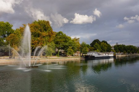 Foto de Hermoso paisaje en un día nublado de otoño en Troyes, Francia, con una fuente con varios chorros en el Canal du Trvois; un barco fluvial está anclado por el agua verde, en un frondoso terraplén - Imagen libre de derechos