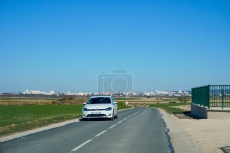 Photo for Les Mesneux, France - March 2022 - A white car drives the D6 Road of Les Mesneux, going away from the city of Reims in the background towards Reims Mountain in the South and crossing the countryside - Royalty Free Image