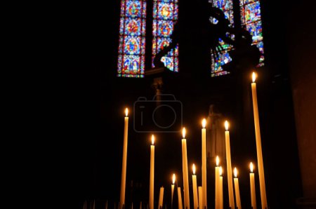 Foto de Reims, Francia - Marzo 2021 - Un conjunto de velas religiosas, brillando en el crepúsculo de una capilla lateral, con vidrieras de colores en el fondo, dentro de la nave de la Catedral de Notre-Dame - Imagen libre de derechos