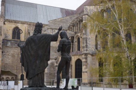 Foto de Reims, Francia - Abril 2021 - Una estatua de bronce de 1996 conmemora el bautismo de Clovis I, rey de los francos, por el obispo Saint-Remi, en 496; en el fondo, el ábside de la Basílica de Saint-Remi - Imagen libre de derechos