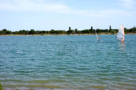 Foto de La orilla y el agua azul del lago y centro de ocio de Rame, en Toulouse, Francia, con windsurfistas disfrutando del día soleado, y árboles en la orilla opuesta - Imagen libre de derechos