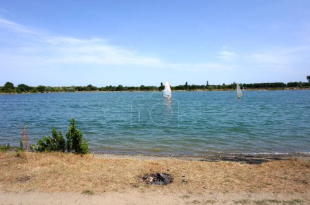 Téléchargez les photos : Le rivage et l'eau bleue du lac et centre de loisirs de Rame, à Toulouse, France, avec des planchistes profitant de la journée ensoleillée, et des arbres sur la rive opposée - en image libre de droit